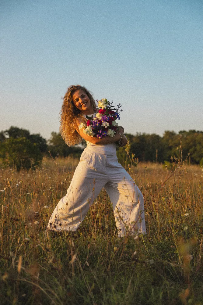 Léa Sucret souriante habillée en blanc tenant un bouquet coloré dans les mains dans un champ au couché du soleil