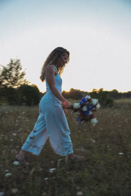 Léa Sucret de profil qui marche habillée en blanc tenant un bouquet coloré dans les mains dans un champ au couché du soleil